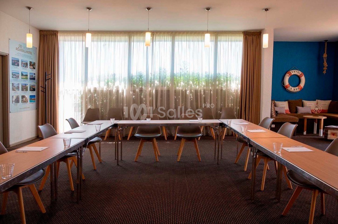 Salle de réunion lumineuse avec tables disposées en U, chaises modernes et décoration nautique en arrière-plan.