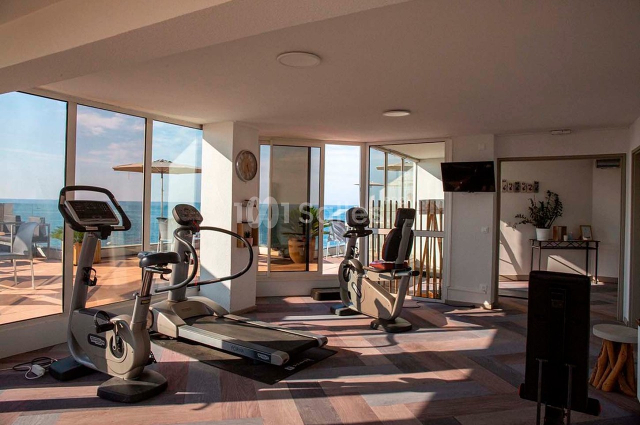 Salle de sport lumineuse avec vélos d'exercice et tapis de course, offrant une vue sur la mer à travers de grandes baies…