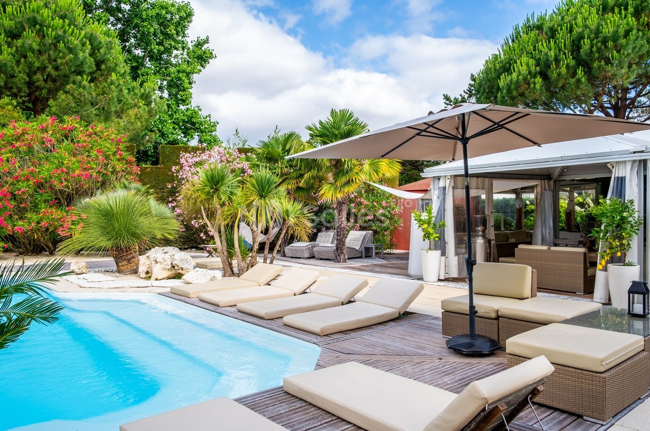 Terrasse avec piscine, transats beiges, parasol et végétation luxuriante sous un ciel partiellement nuageux.