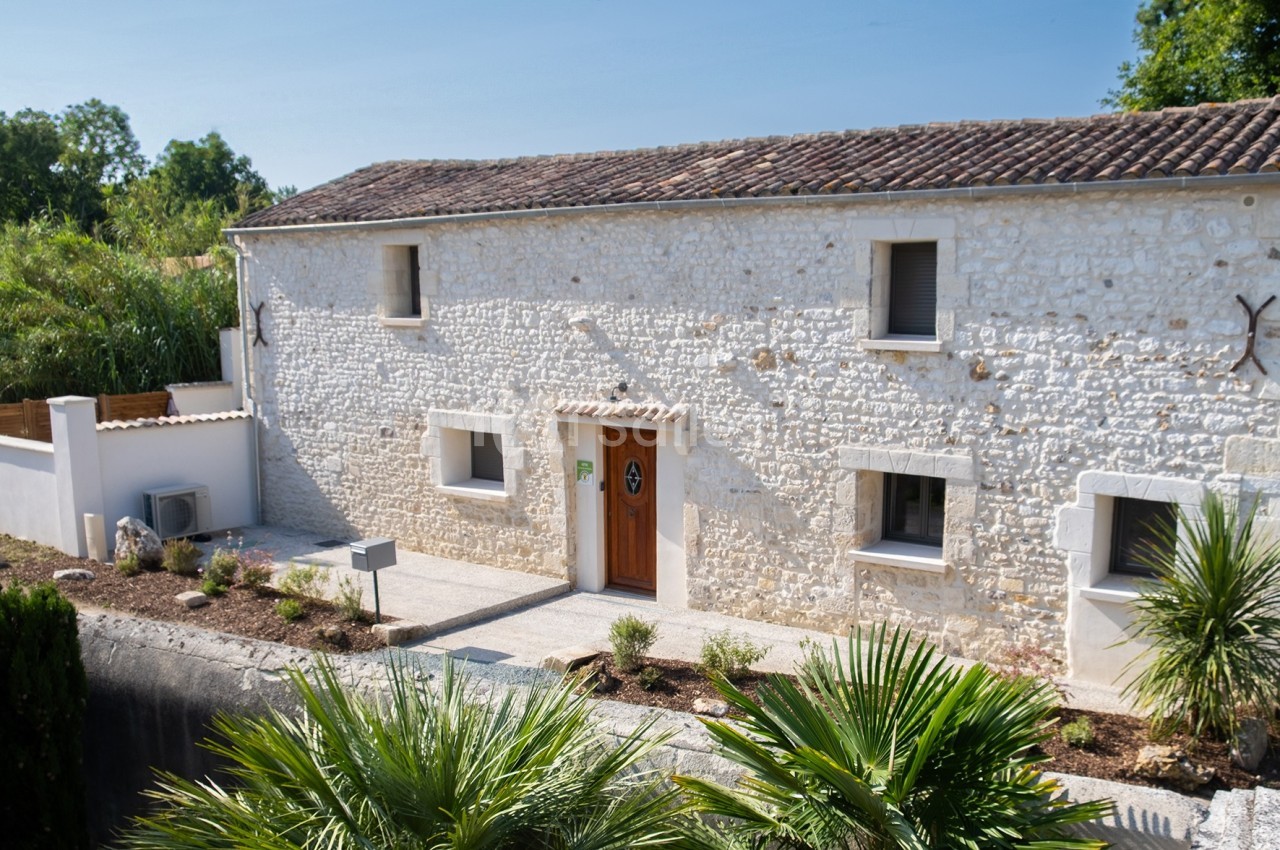 Façade d'une maison en pierre blanche avec toit en tuiles, entourée de végétation et d'un petit jardin.