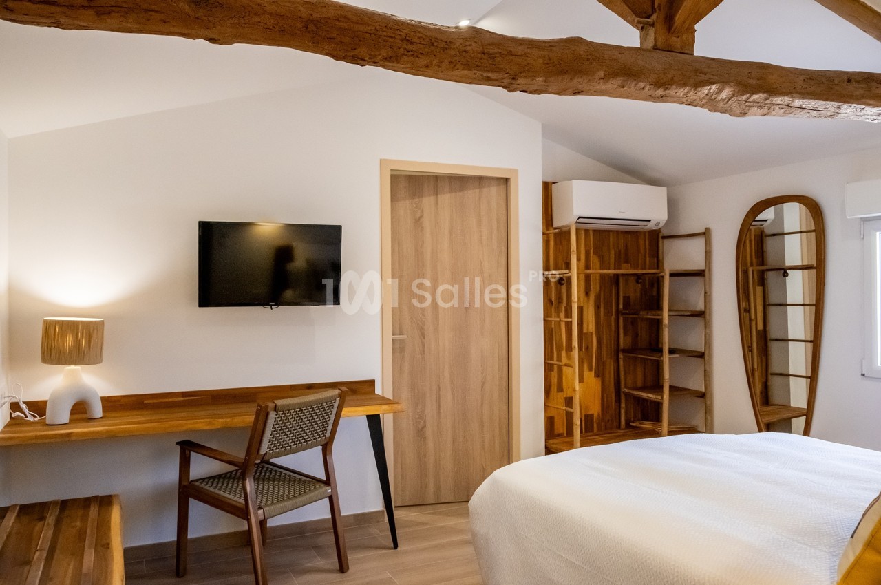 Chambre lumineuse avec poutres apparentes, lit double, bureau en bois, télévision murale et étagères ouvertes.