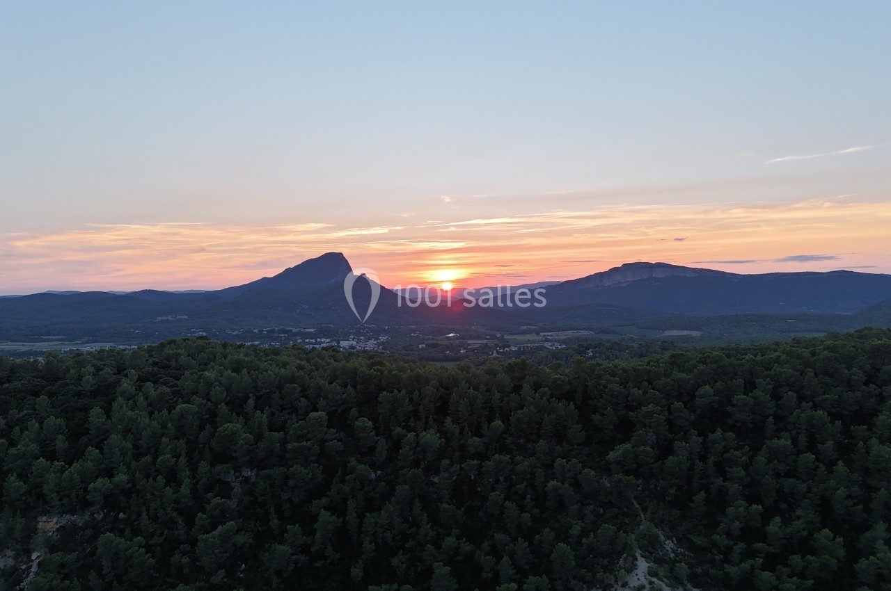 Coucher de soleil derrière des montagnes, avec une forêt dense au premier plan et un ciel aux teintes orangées.