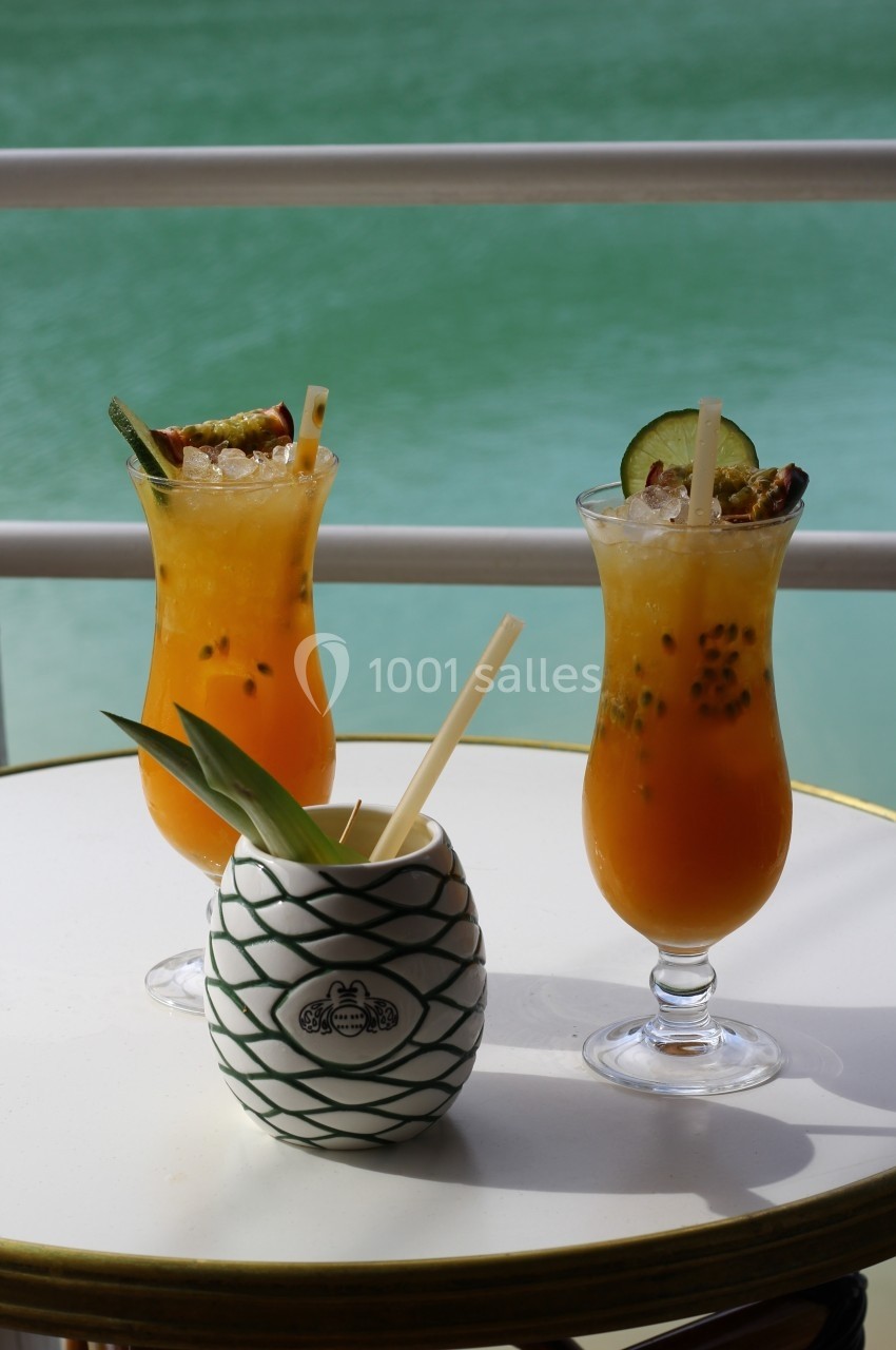 Deux cocktails tropicaux et un verre décoratif sur une table blanche, avec vue sur une eau turquoise en arrière-plan.