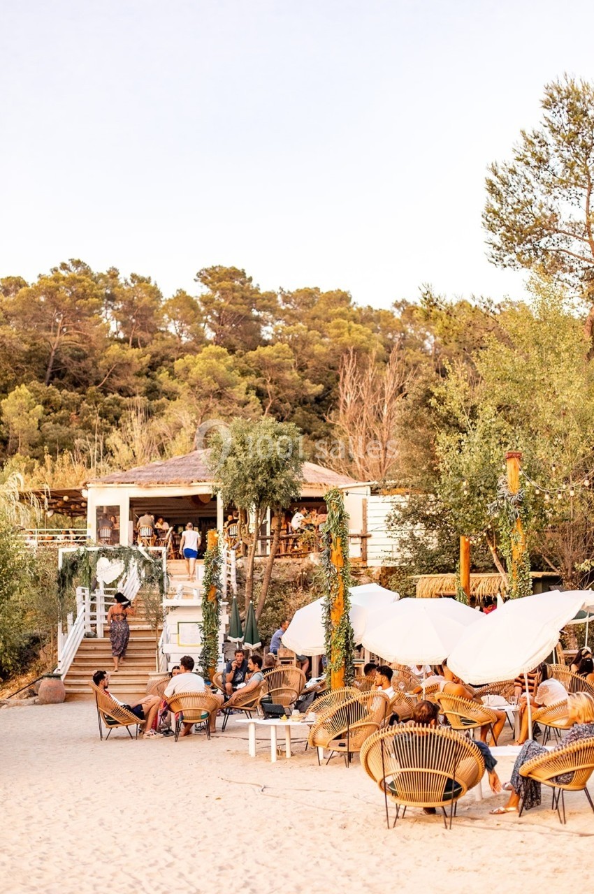 Terrasse d'un restaurant en bord de plage avec des parasols blancs, des chaises en rotin et une végétation environnante.