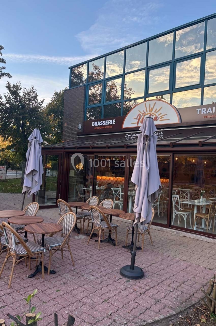 Terrasse d'une brasserie avec tables et chaises en osier, parasols fermés, devant une façade vitrée au coucher du soleil.