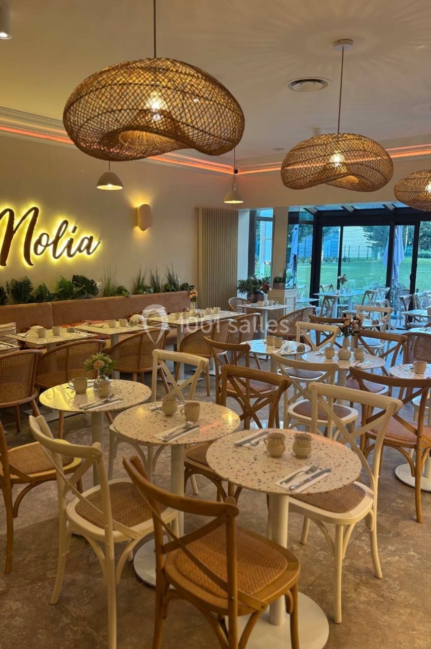 Salle de restaurant lumineuse avec chaises en bois, tables rondes et luminaires suspendus en osier.