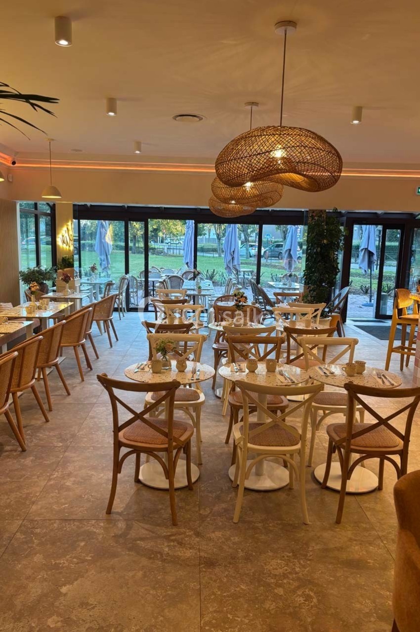 Salle de restaurant lumineuse avec tables en bois clair, chaises assorties et vue sur une terrasse extérieure arborée.