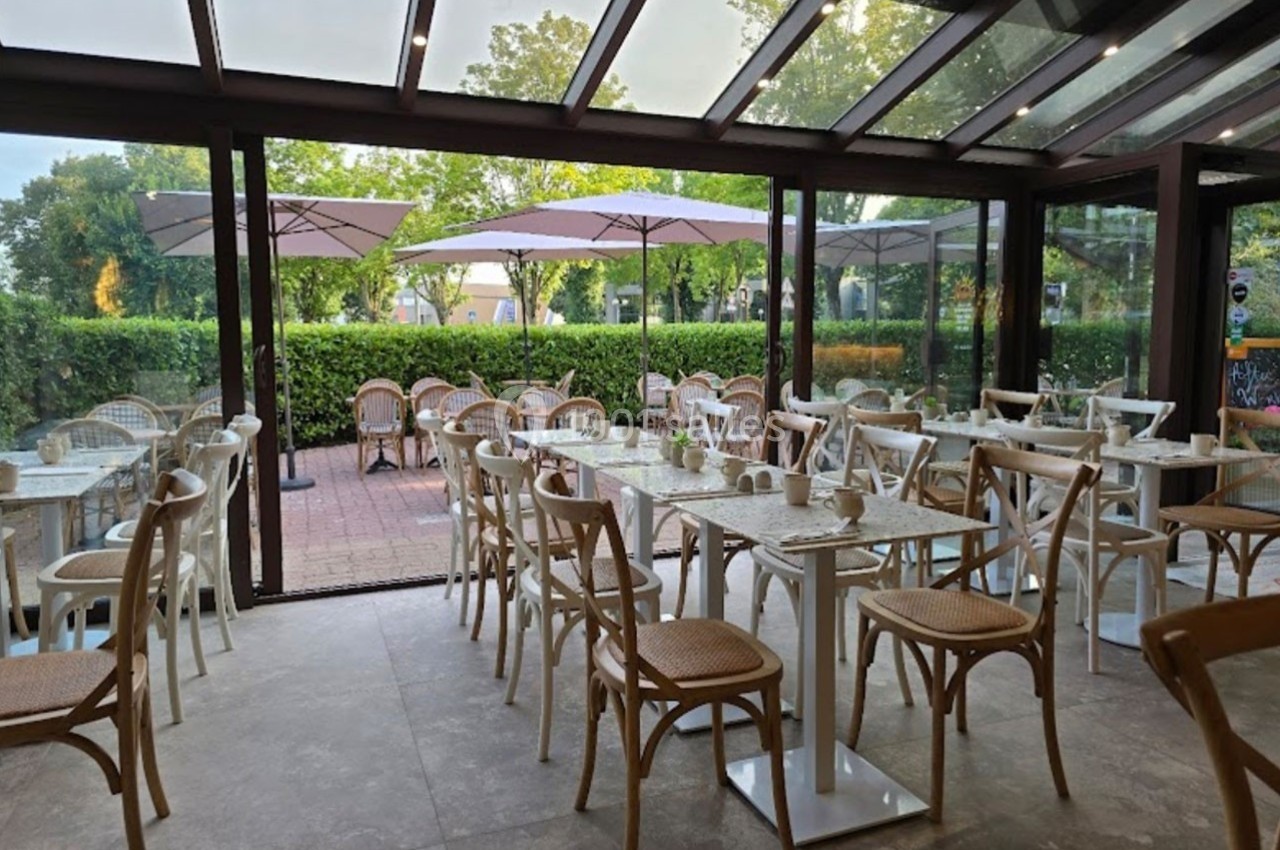 Salle de restaurant lumineuse avec des tables et chaises en bois clair, donnant sur une terrasse avec parasols et verdure.