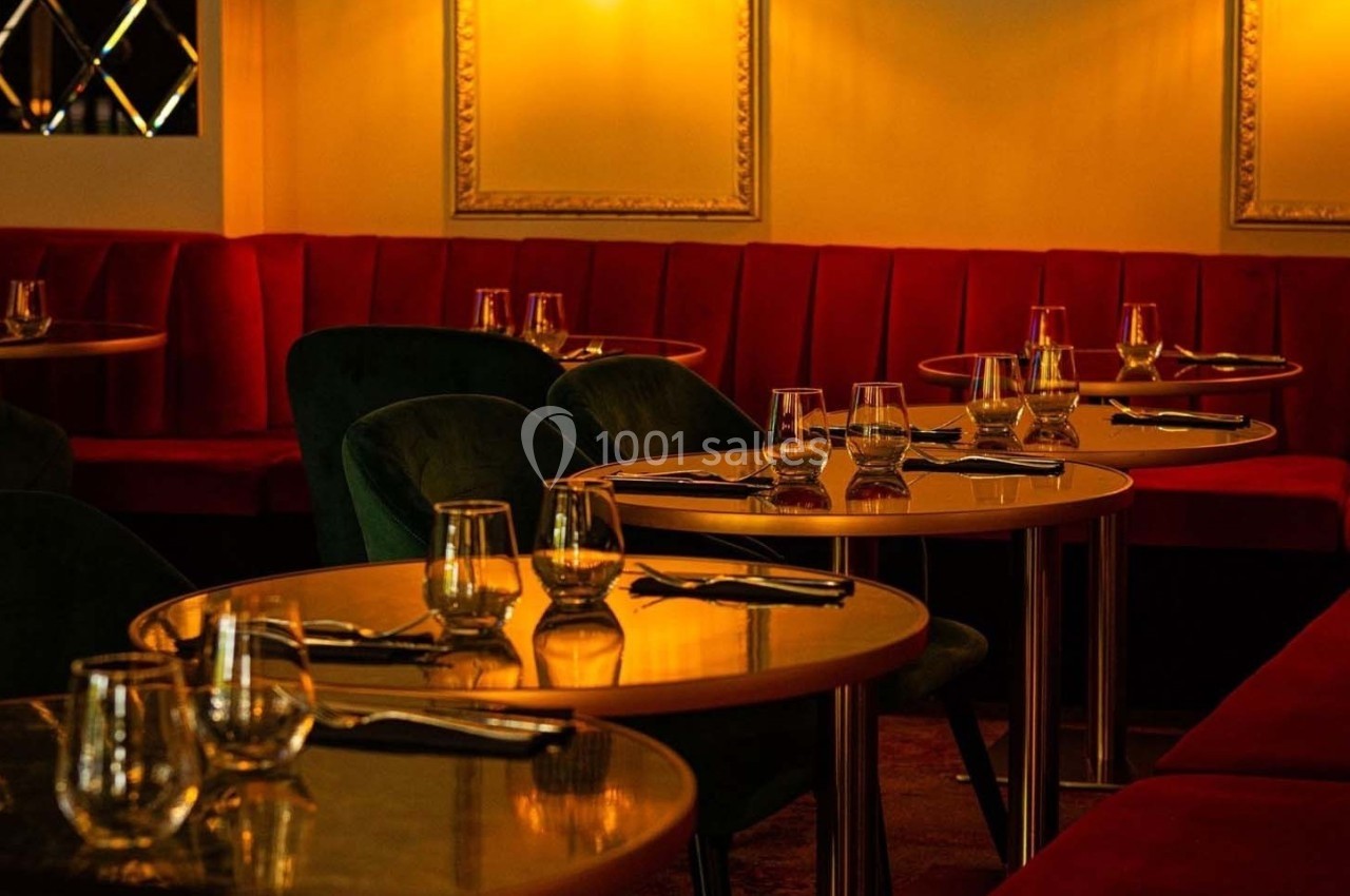 Salle de restaurant avec tables rondes dorées, chaises vertes et banquettes rouges dans une ambiance tamisée.