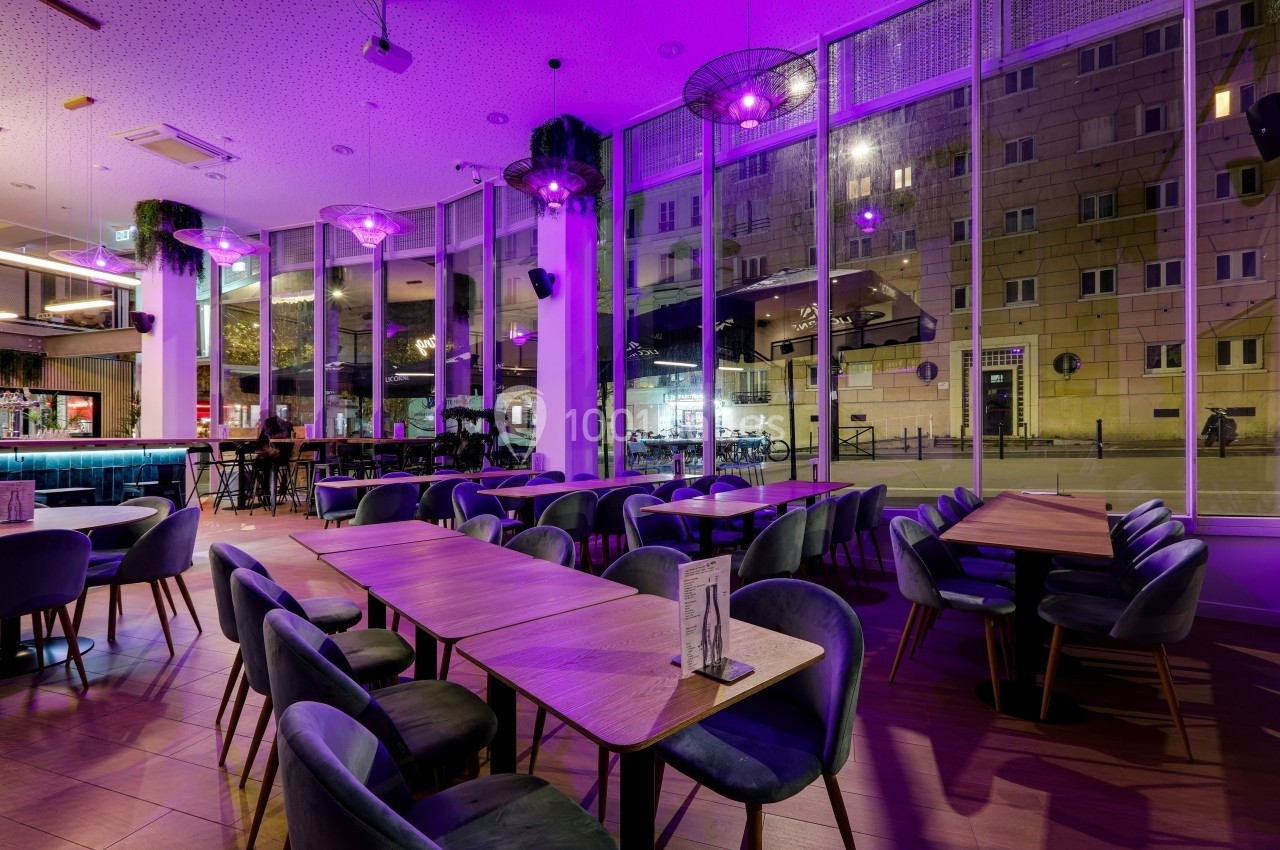 Salle de restaurant moderne avec éclairage violet, grandes baies vitrées et tables en bois entourées de chaises rembourrées.