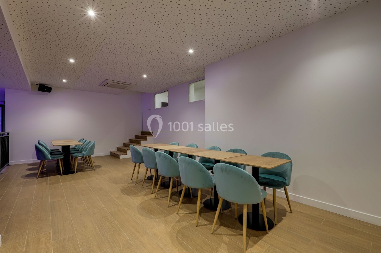 Salle moderne avec tables en bois, chaises bleu-vert, sol en parquet et plafond perforé éclairé par des spots.