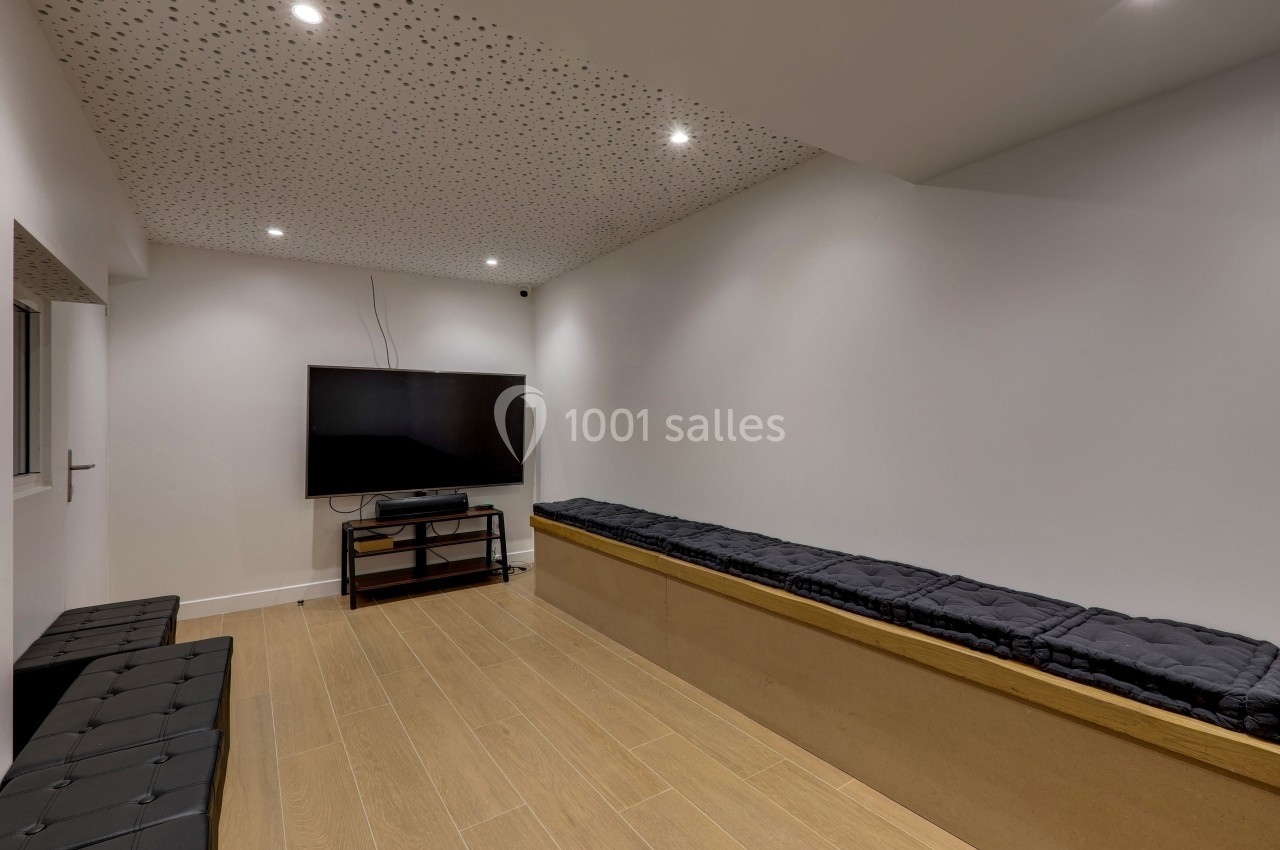 Salle avec un écran plat fixé sur un meuble, bancs rembourrés, sol en bois clair et plafond perforé avec spots intégrés.