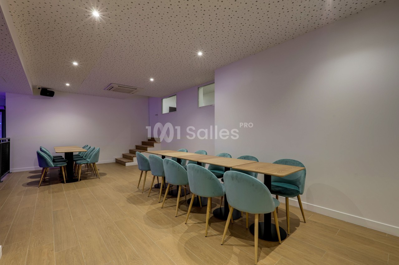 Salle moderne avec tables en bois, chaises bleu-vert, sol en parquet clair et plafond perforé éclairé par des spots.