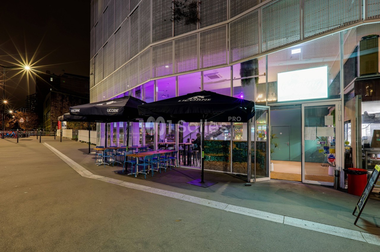 Terrasse extérieure d'un café-restaurant éclairée la nuit, avec tables, chaises et parasols noirs.