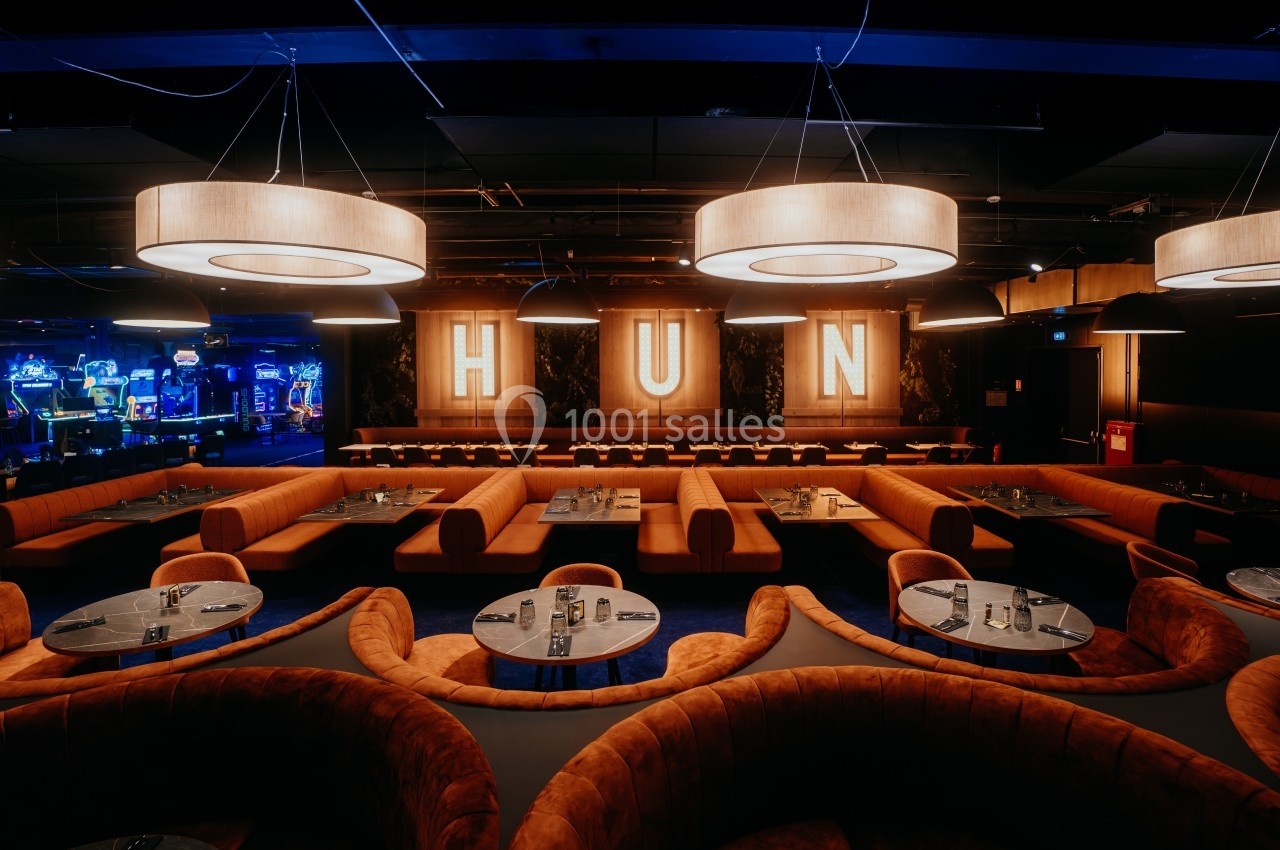 Salle de restaurant moderne avec banquettes orange, tables rondes et grandes lettres lumineuses formant ’HUN’ en arrière…
