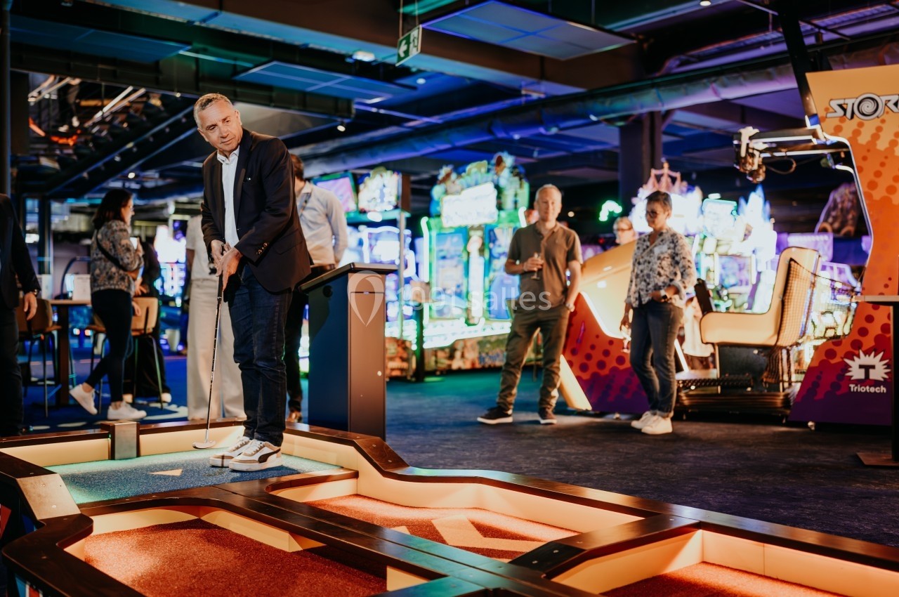 Un homme joue au mini-golf dans un espace intérieur lumineux avec des jeux d'arcade en arrière-plan.