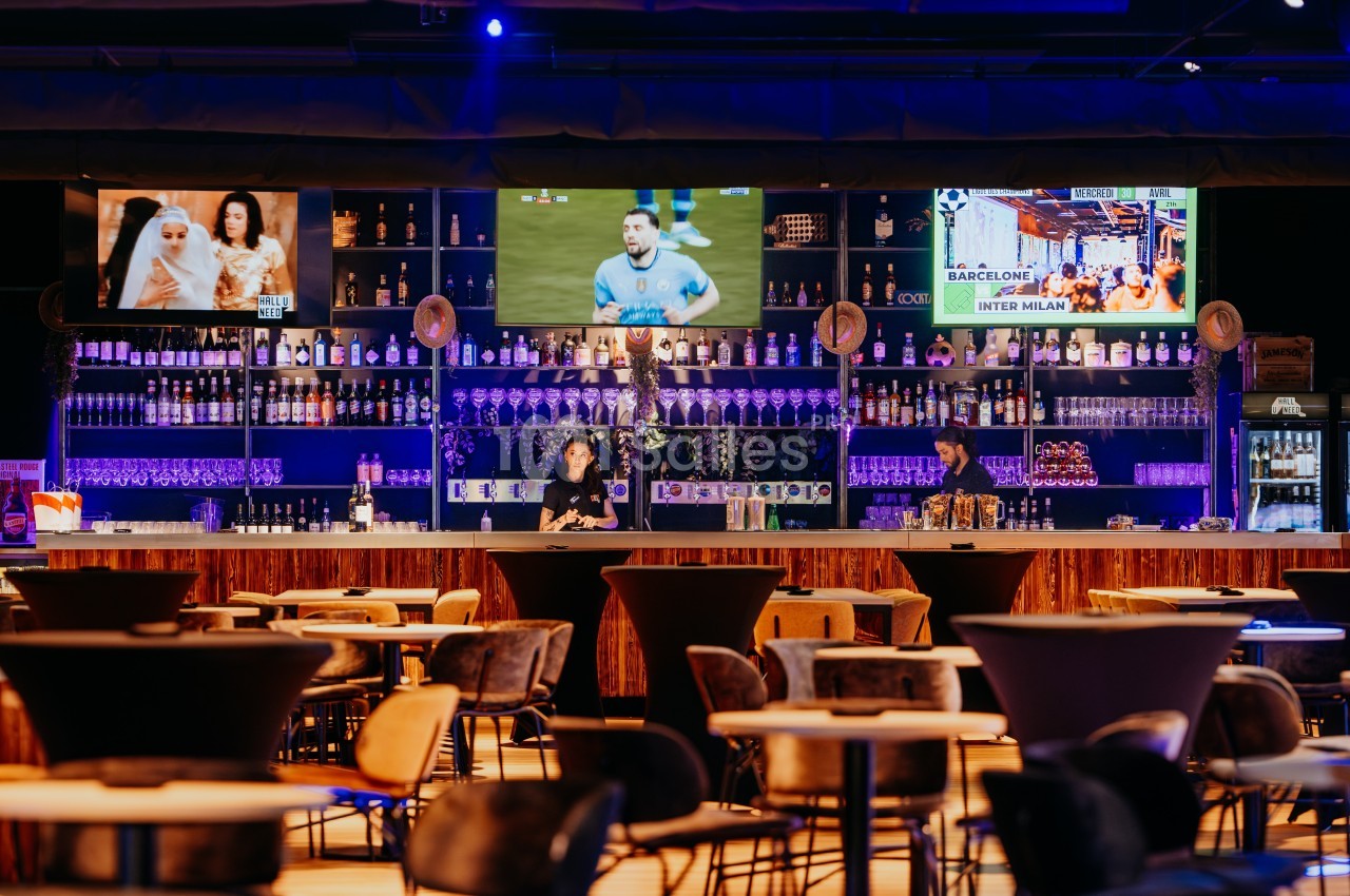 Bar moderne avec étagères remplies de bouteilles, écrans diffusant des matchs et tables disposées en salle.