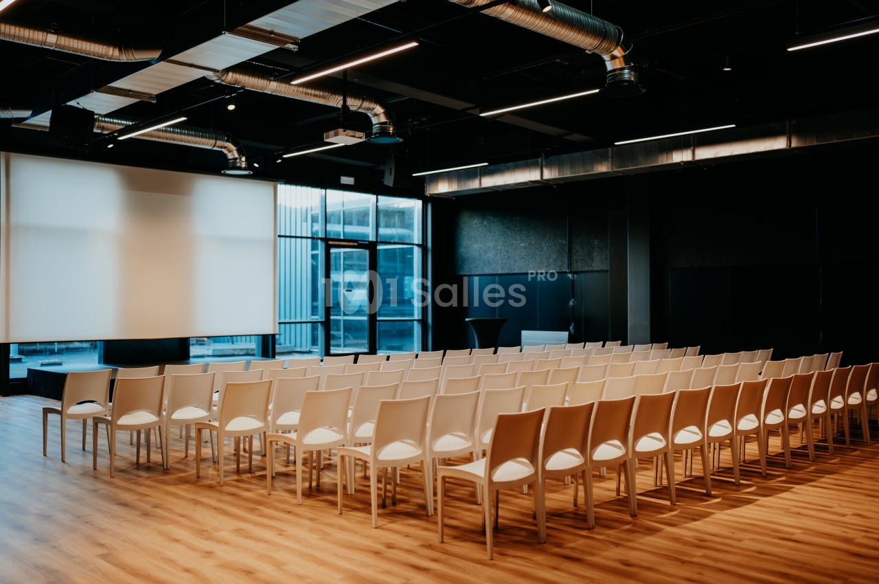 Salle de conférence moderne avec des rangées de chaises blanches alignées face à un écran de projection.