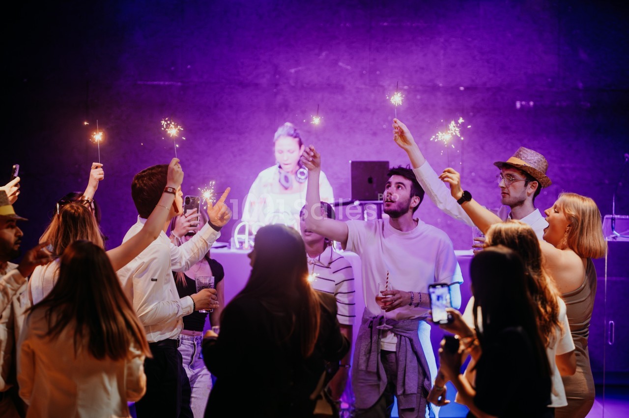 Groupe de personnes célébrant avec des cierges magiques devant un DJ sur une scène éclairée par des lumières violettes.