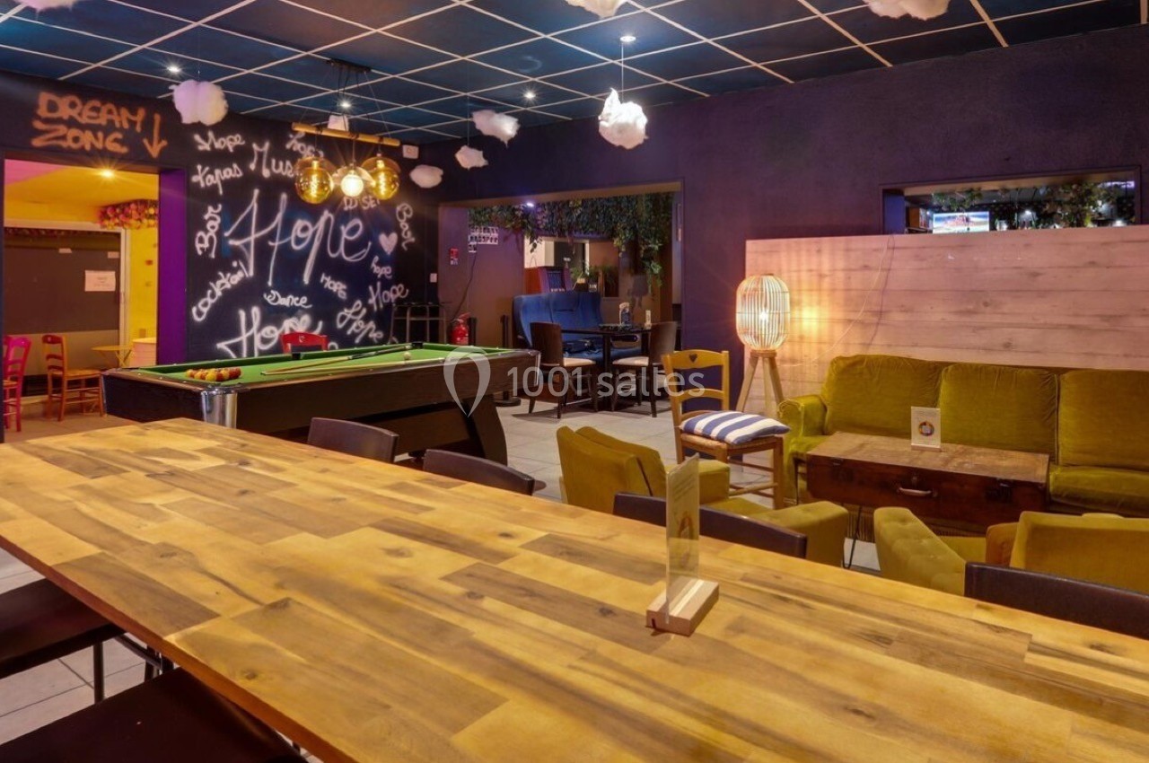 Salle conviviale avec table en bois, billard, canapés colorés et décorations murales lumineuses.