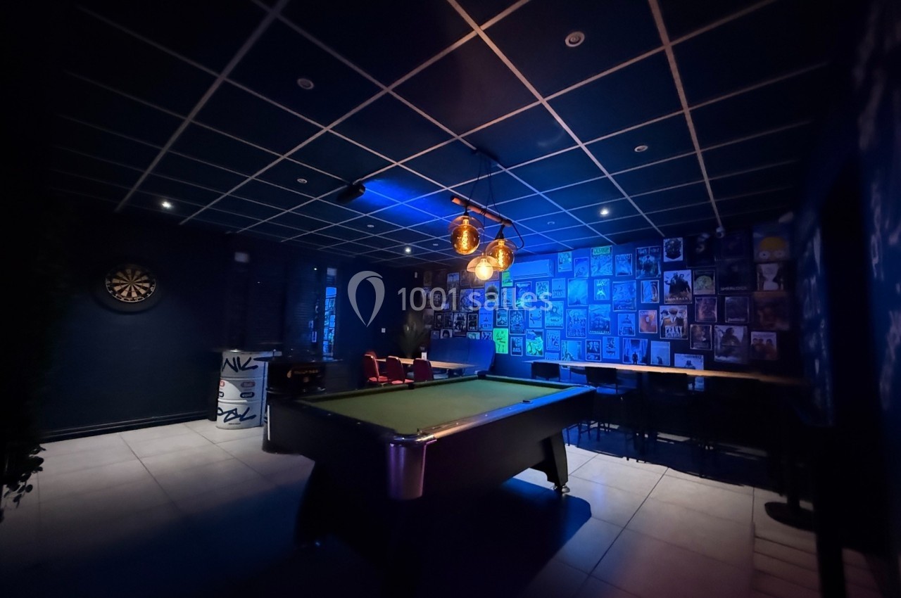 Salle sombre avec une table de billard éclairée, un mur décoré de posters et une cible de fléchettes au fond.