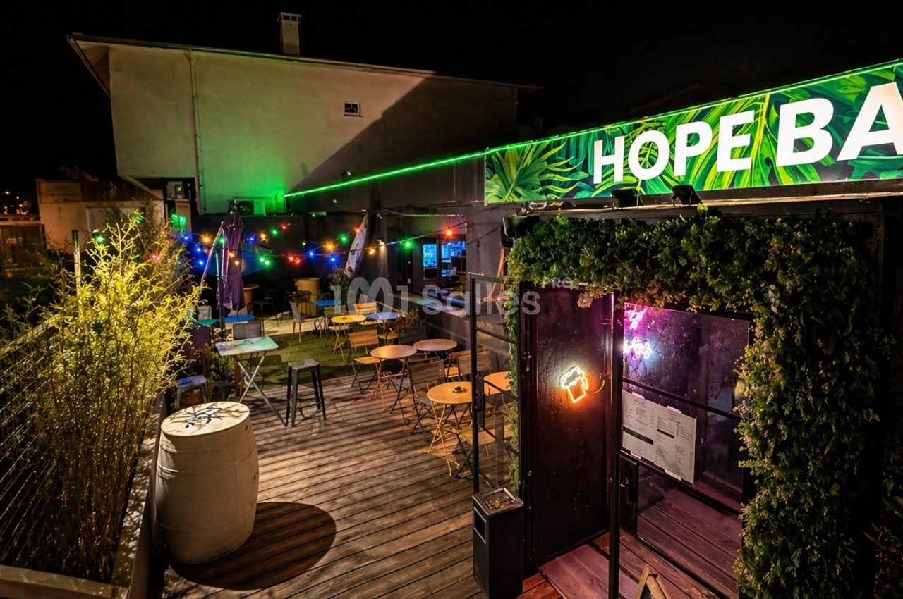 Terrasse extérieure d'un bar éclairée de guirlandes lumineuses colorées, avec tables et chaises disposées.