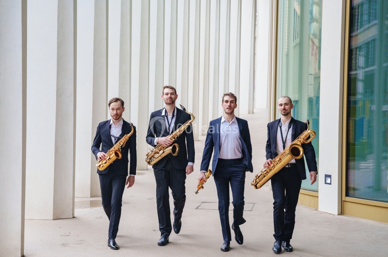 Quatre hommes en costume marchant côte à côte, chacun tenant un saxophone, devant un bâtiment moderne.