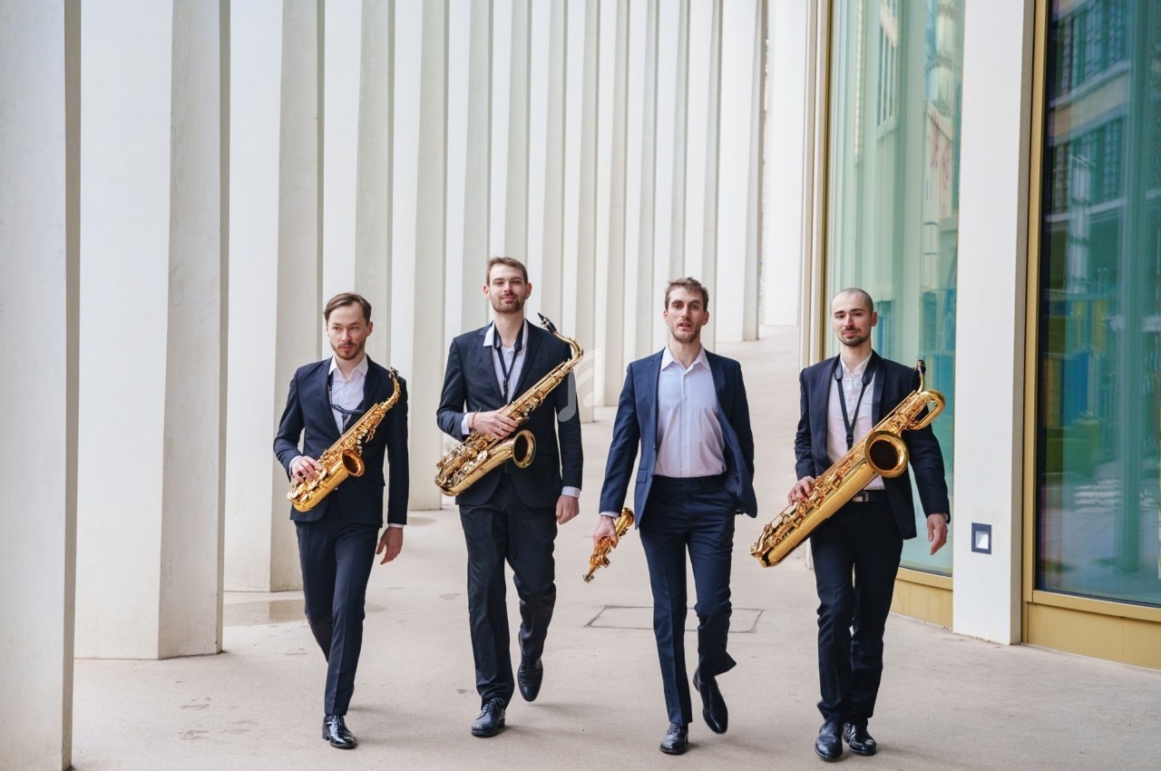 Quatre hommes en costume marchant côte à côte, chacun tenant un saxophone, devant un bâtiment moderne.