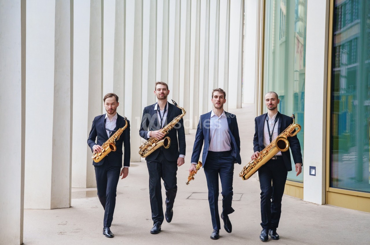 Quatre hommes en costume marchent côte à côte en tenant des saxophones devant un bâtiment moderne.