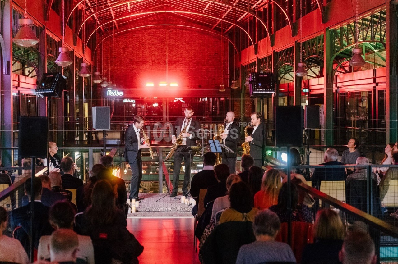 Un groupe de musiciens joue devant un public assis dans un espace intérieur illuminé par des lumières rouges.