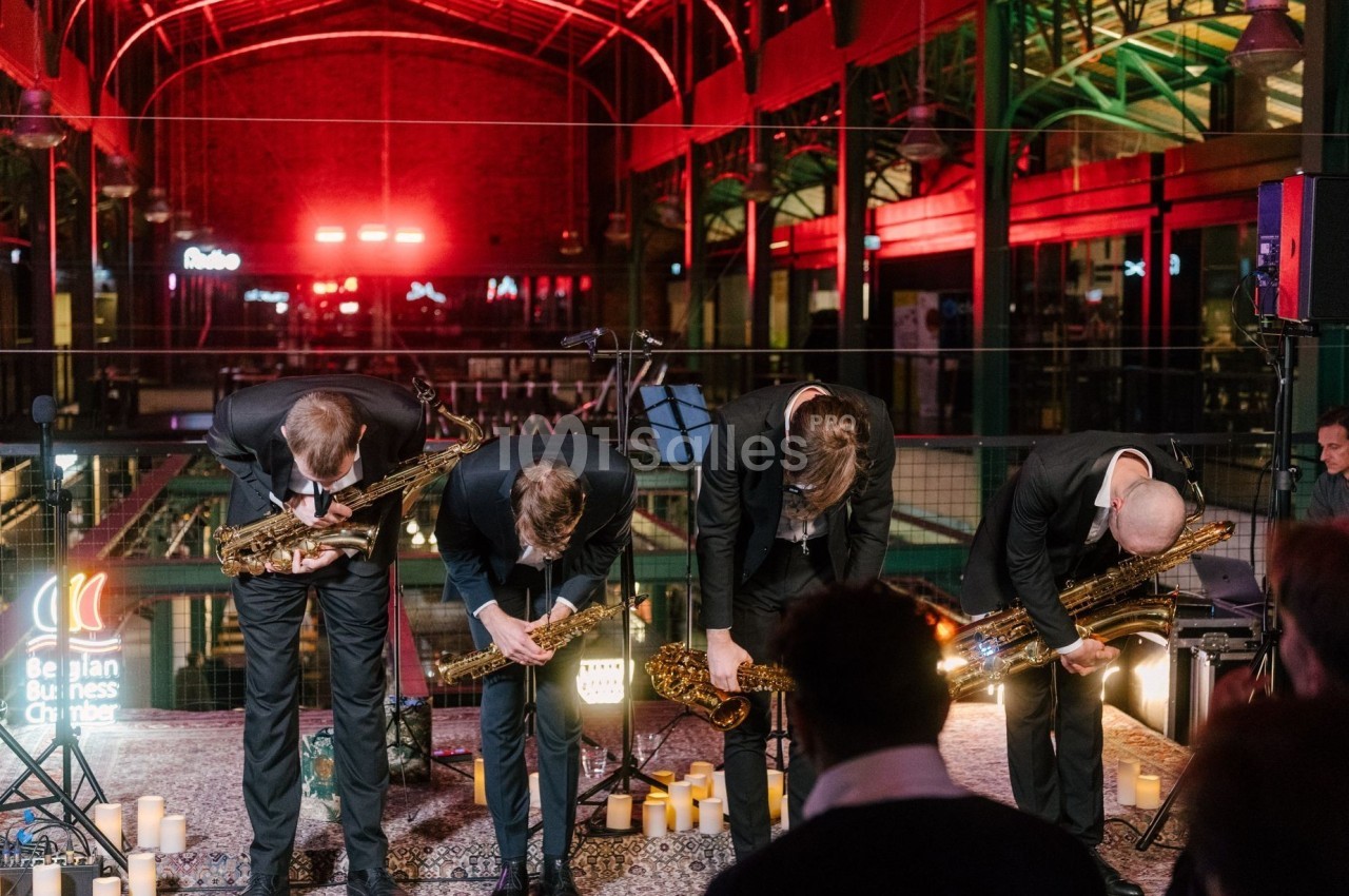 Quatre musiciens en costume s'inclinent avec leurs saxophones sur une scène éclairée, entourée de bougies.