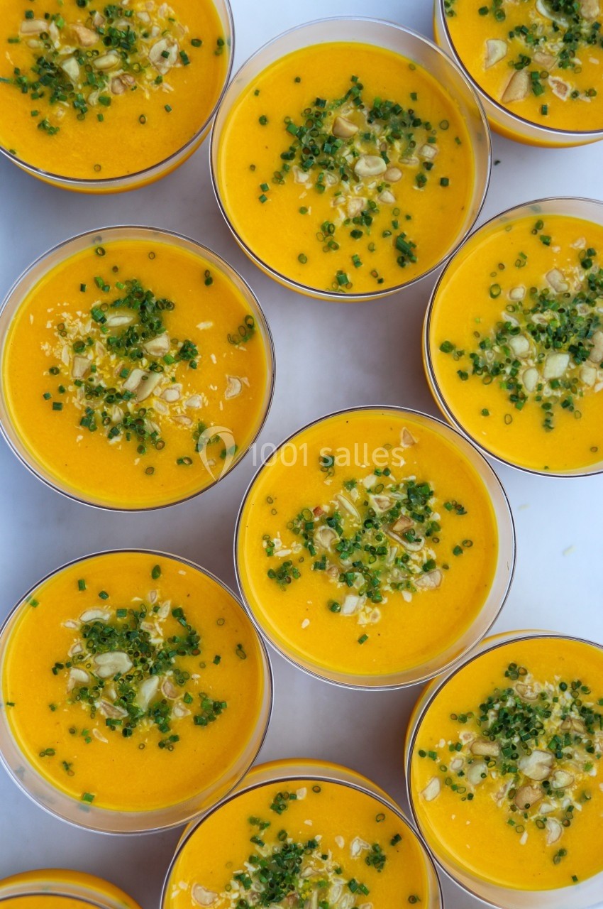Petits bols de soupe orange garnis de ciboulette hachée, graines et morceaux de légumes sur une surface blanche.