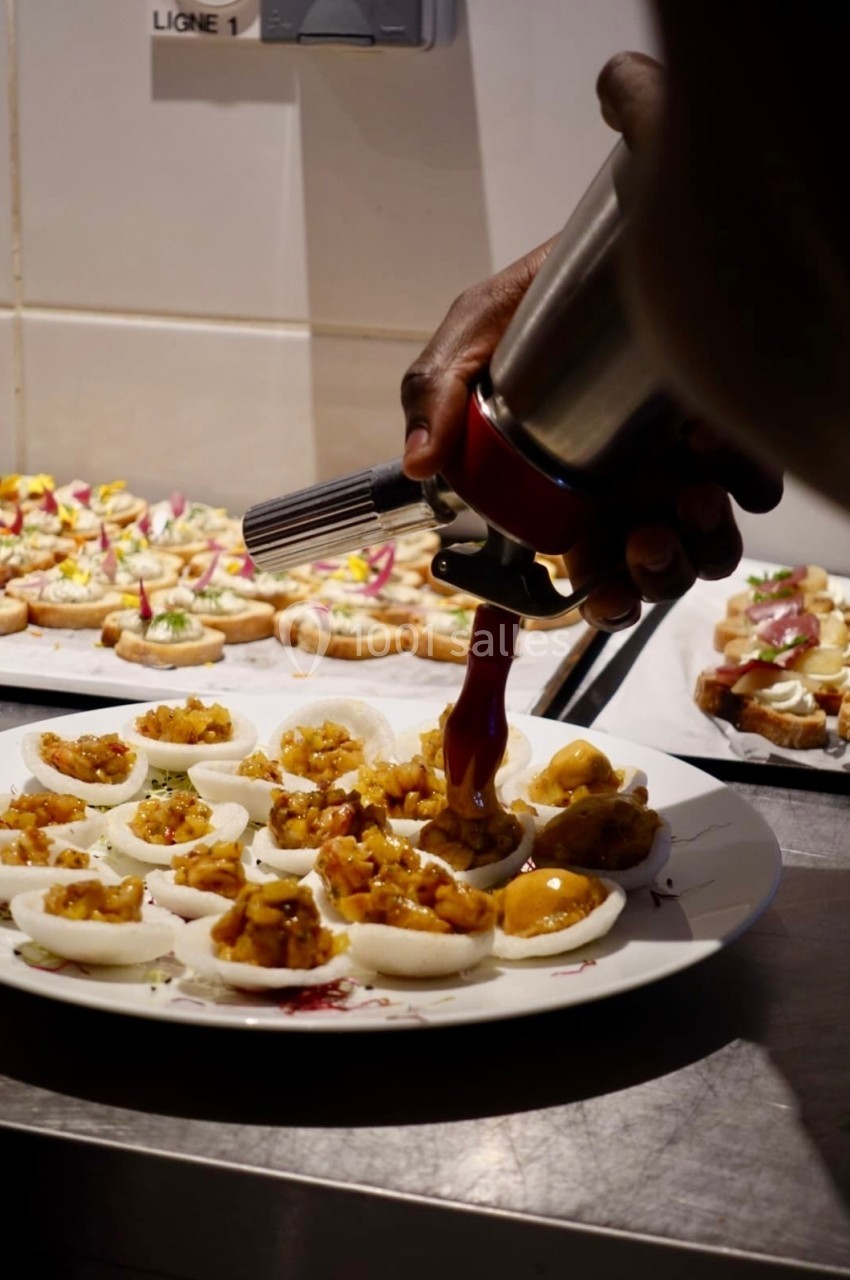 Une main garnit des bouchées apéritives avec une poche à douille dans une cuisine professionnelle.
