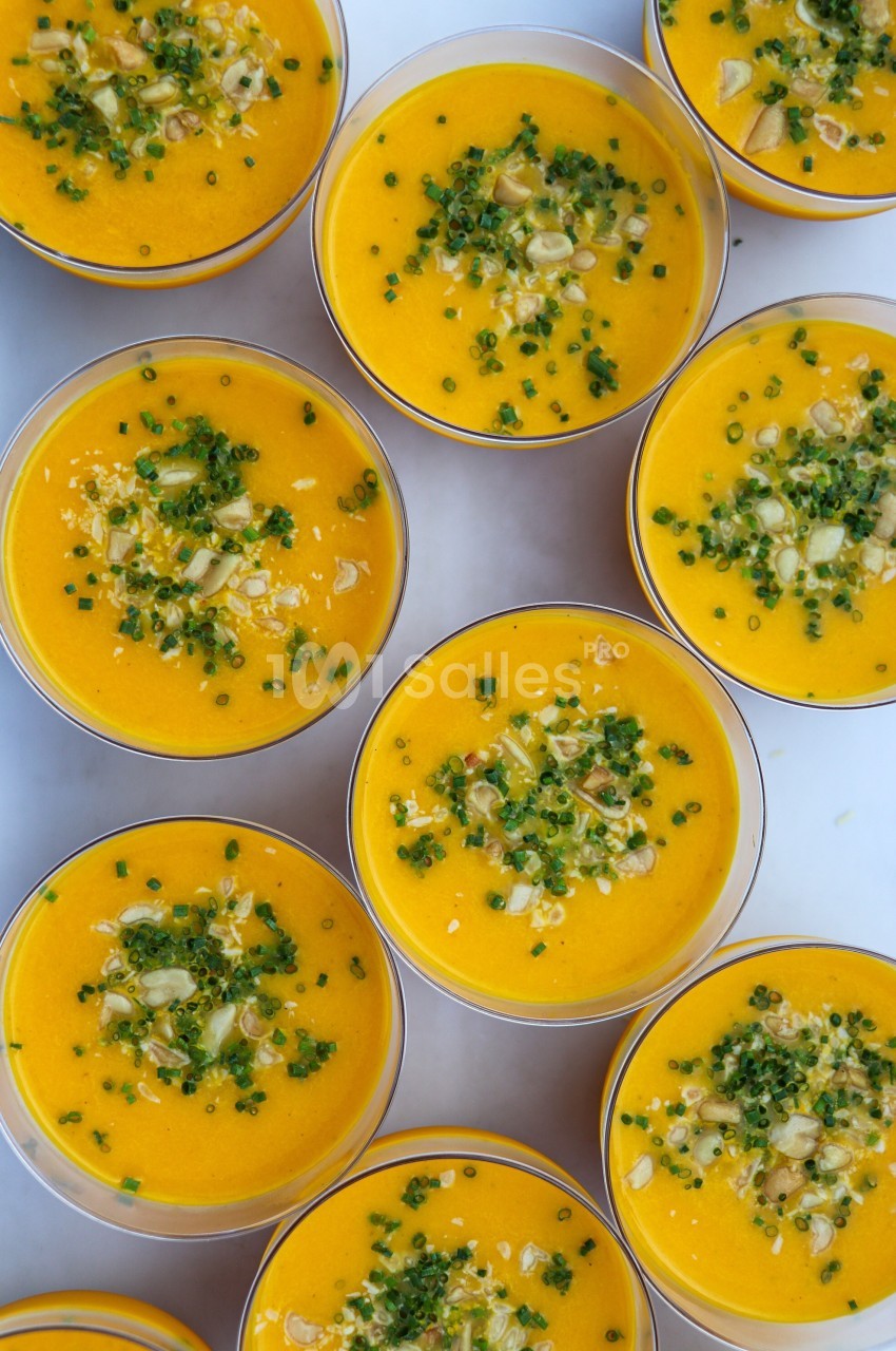 Petits bols de soupe orange garnis de ciboulette hachée, graines et morceaux de légumes sur une surface blanche.