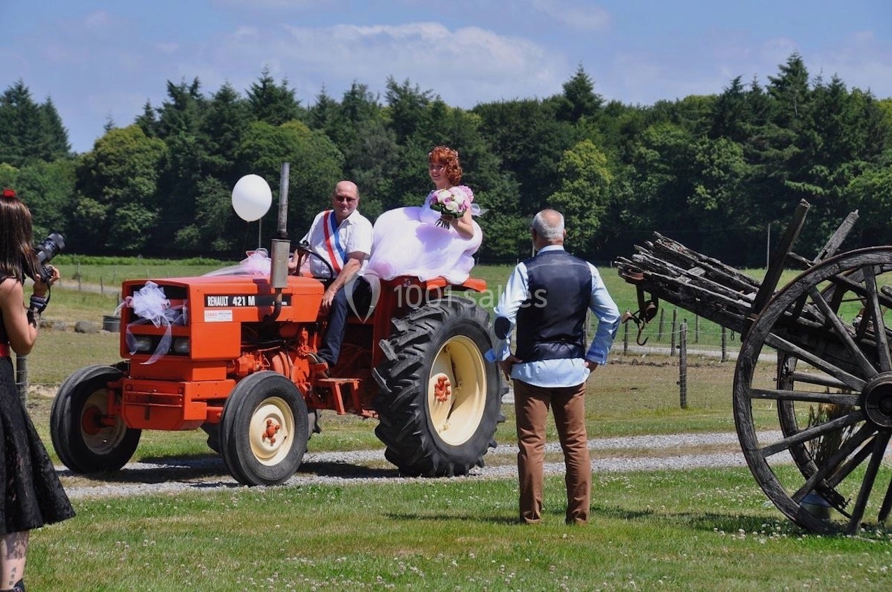 Une mariée en robe blanche est assise sur un tracteur rouge décoré, entourée de personnes dans un cadre champêtre.