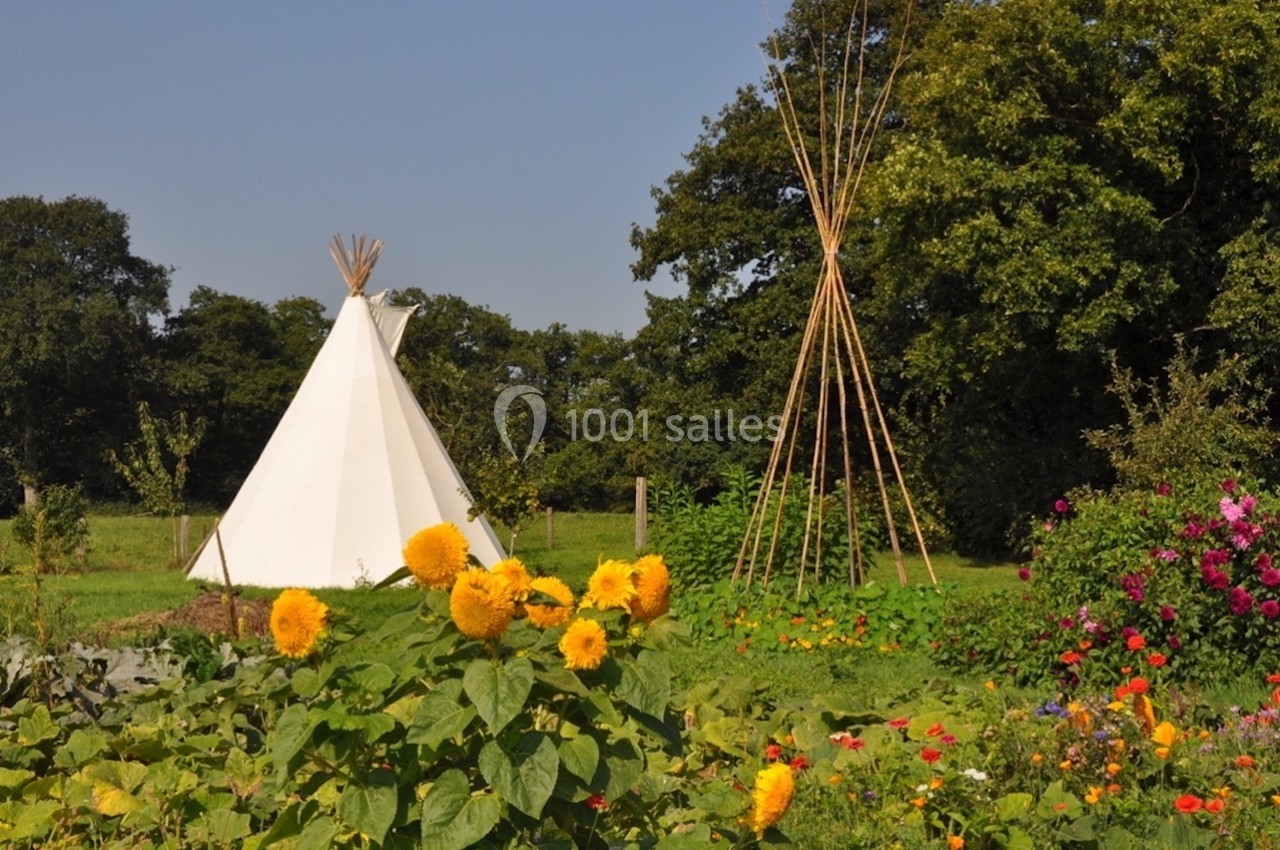 Un tipi blanc et une structure en bois dans un jardin fleuri avec des tournesols, entourés d'arbres et de verdure.