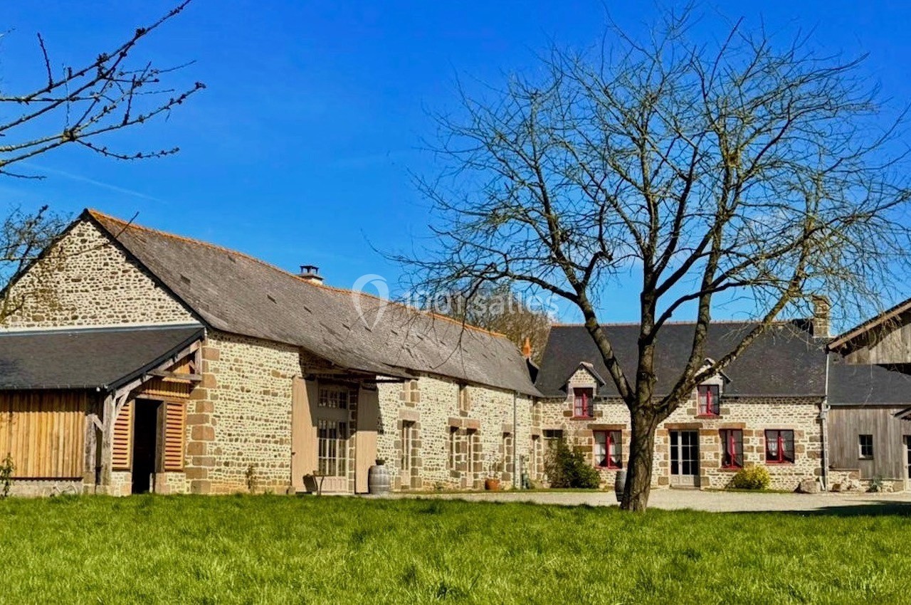 Ferme en pierre avec toit en ardoise, entourée d'herbe et d'un arbre, sous un ciel bleu dégagé.