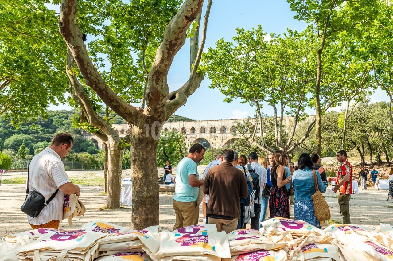 Un groupe de personnes rassemblées sous des arbres près d'un pont historique, avec des sacs en tissu au premier plan.