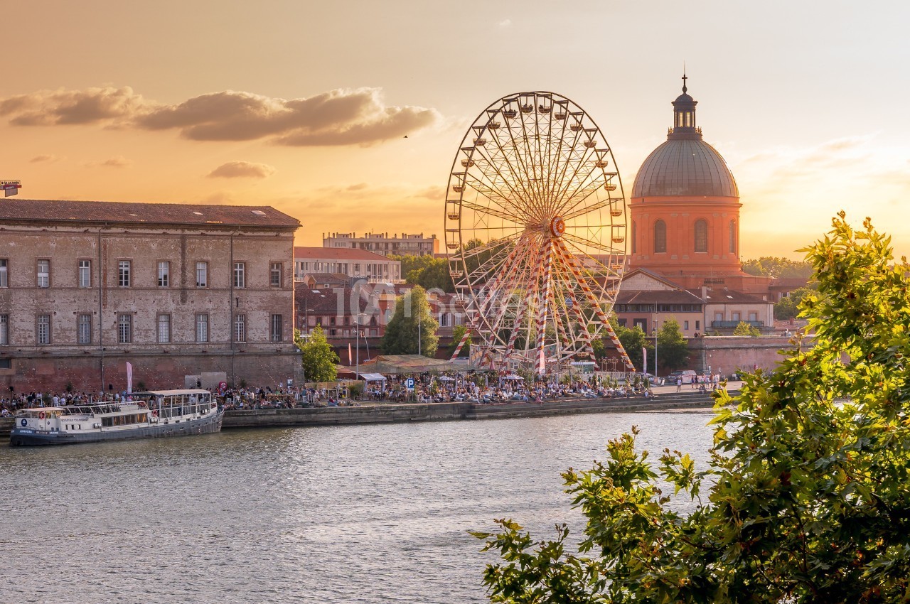 Vue sur une grande roue, un dôme et des bâtiments historiques au bord d'une rivière au coucher du soleil.