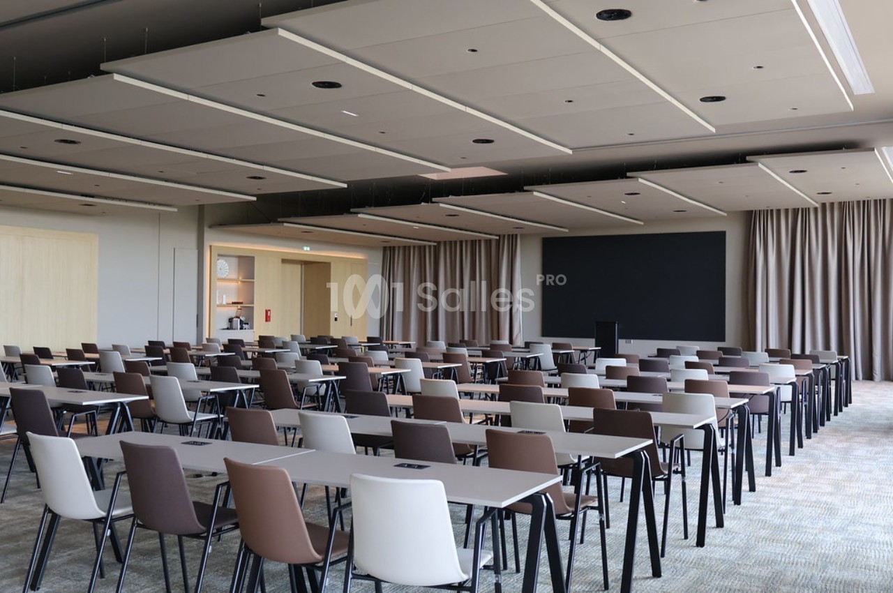 Salle de conférence moderne avec des rangées de tables et de chaises alignées face à un écran noir.