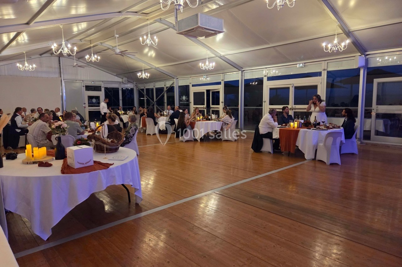 Salle de réception éclairée avec des invités assis à des tables décorées, ambiance chaleureuse et conviviale.