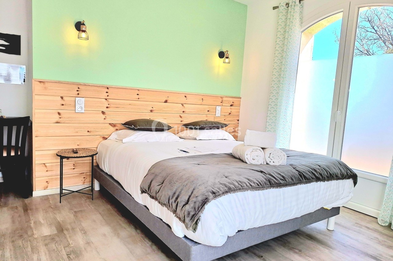 Chambre lumineuse avec lit double, tête de lit en bois, murs verts, parquet clair et grande fenêtre avec rideaux.