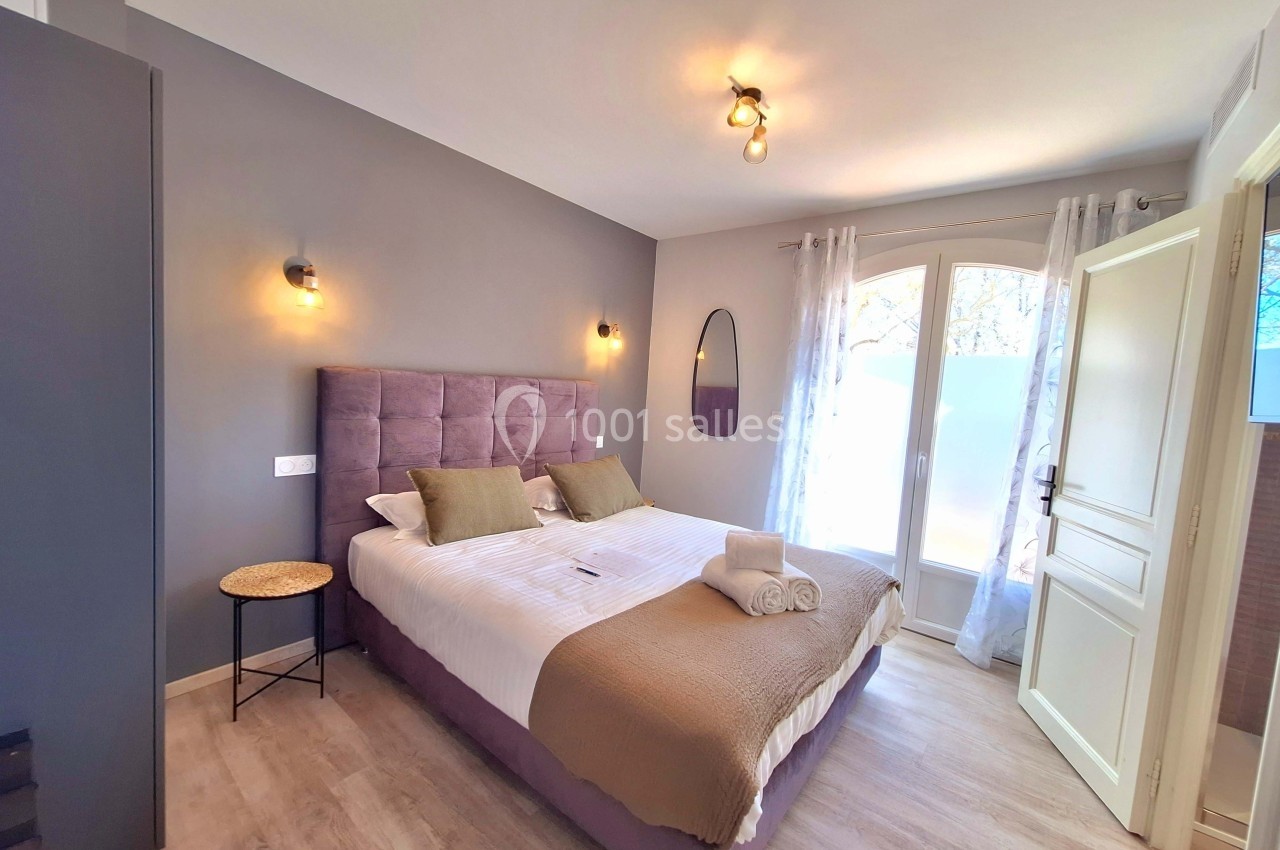 Chambre lumineuse avec lit double, murs gris, éclairage mural, table d'appoint et grande fenêtre avec rideaux.