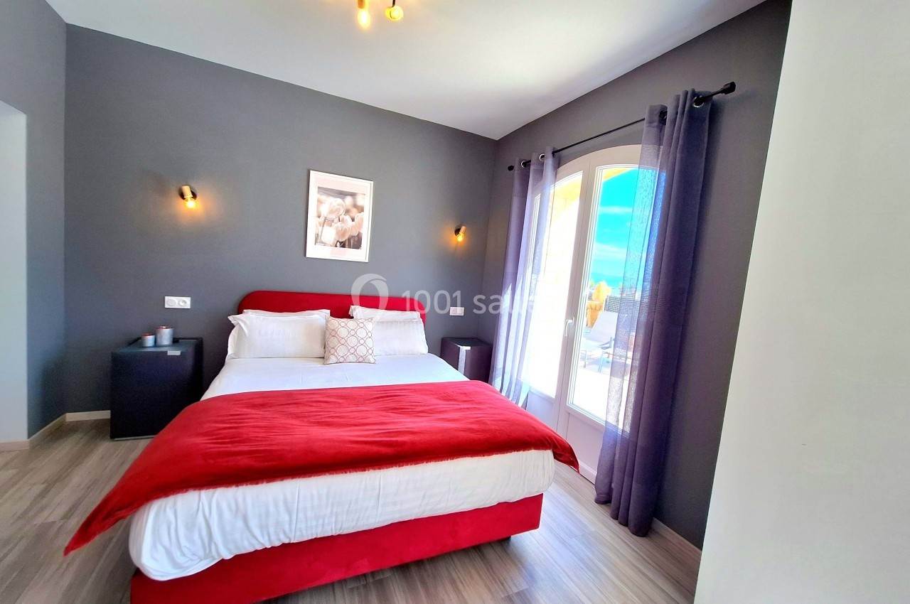 Chambre lumineuse avec lit double, murs gris, parquet clair, rideaux violets et accès à un balcon ensoleillé.