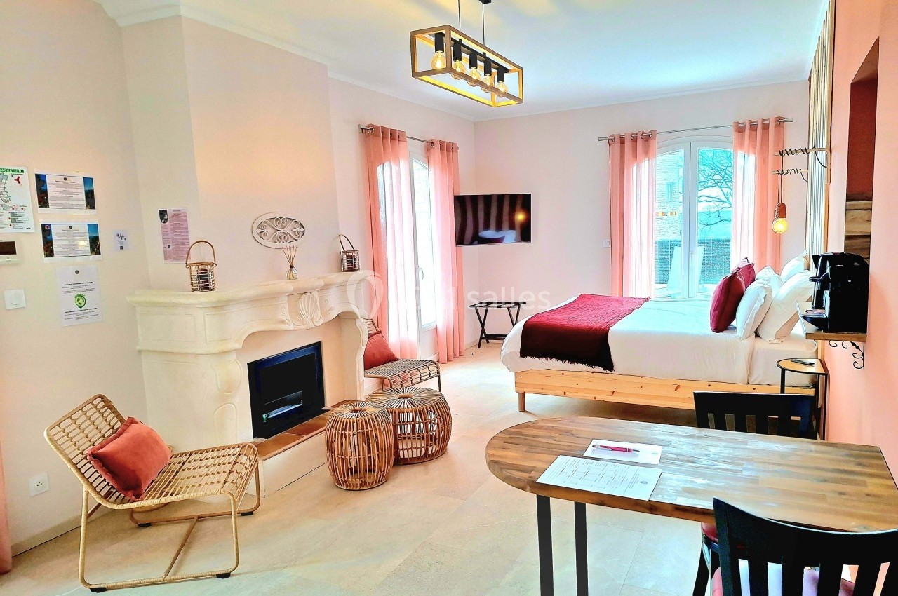 Chambre lumineuse avec lit double, cheminée, table en bois, fauteuils en rotin et grandes fenêtres avec rideaux roses.