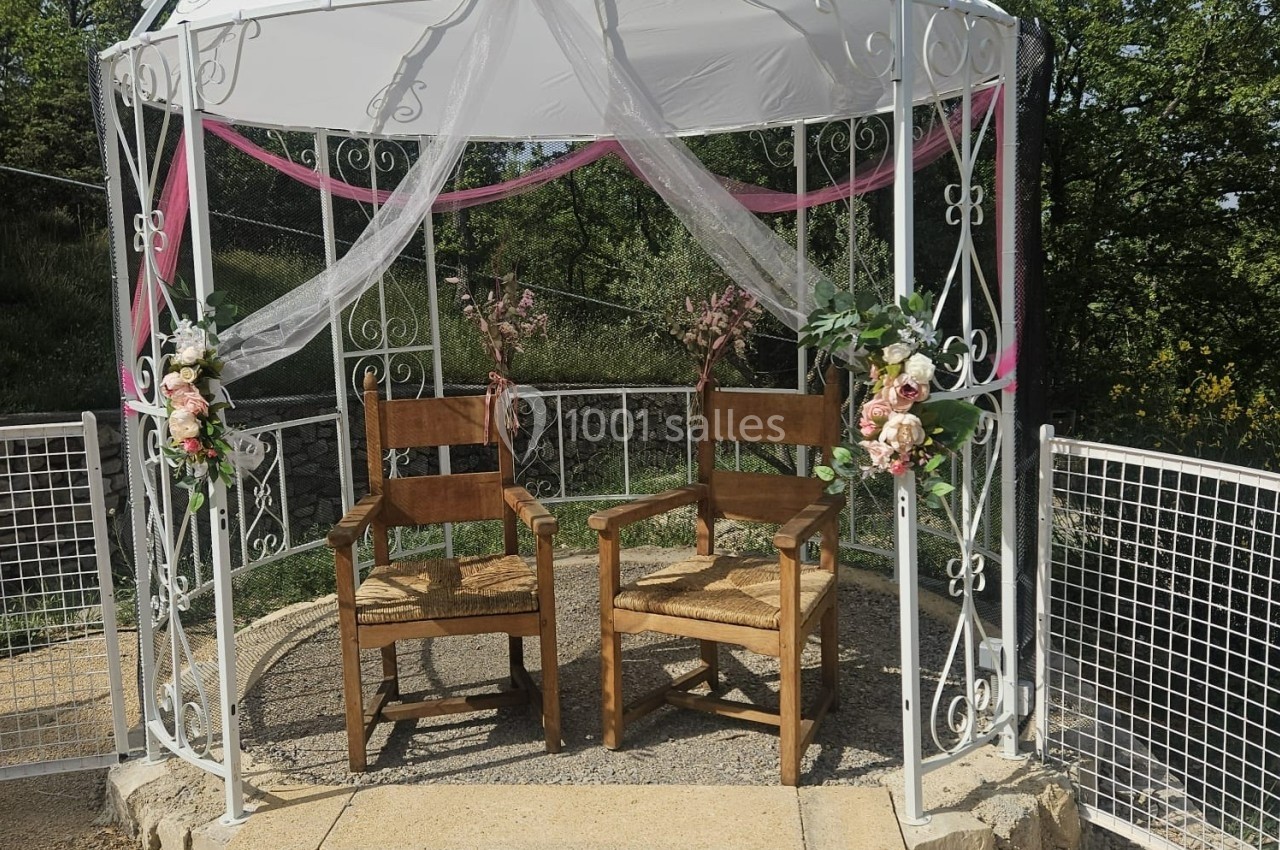 Deux chaises en bois sous une tonnelle blanche décorée de voilages et de fleurs, en extérieur.
