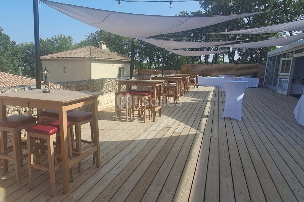 Terrasse en bois avec tables hautes, chaises rouges et voiles d'ombrage, entourée de bâtiments et d'arbres.