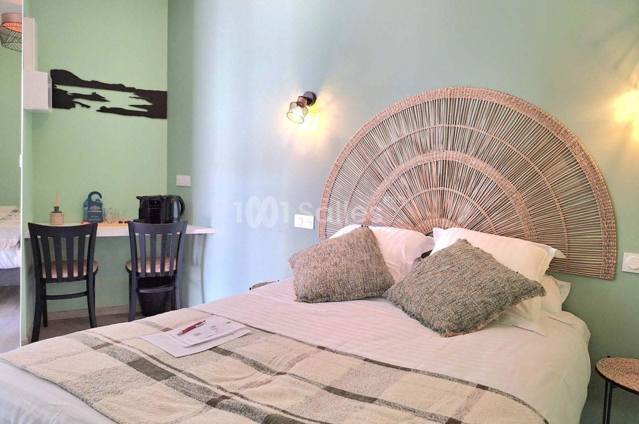 Chambre lumineuse avec un lit double, tête de lit en rotin, bureau avec chaises et mur peint en vert clair.