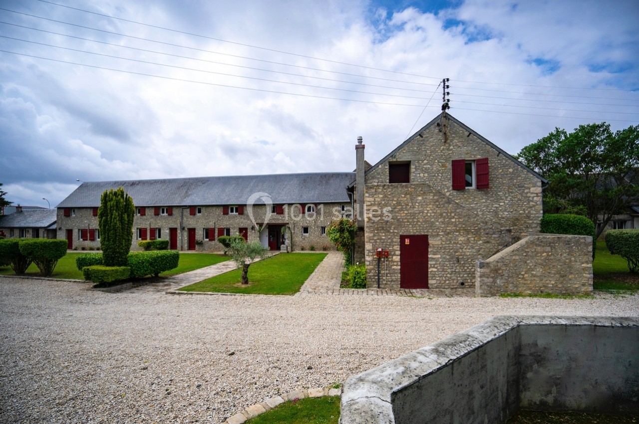 Bâtiment en pierre avec volets rouges, entouré de pelouses et d'arbustes, sous un ciel nuageux.