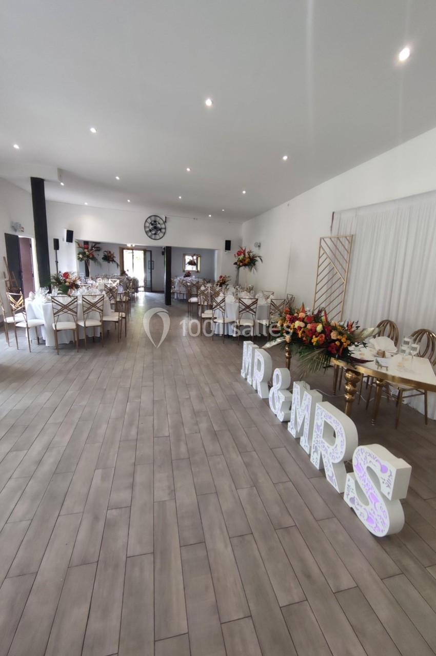 Salle de réception décorée pour un mariage, avec tables dressées, compositions florales et lettres lumineuses ’MR & MRS’.